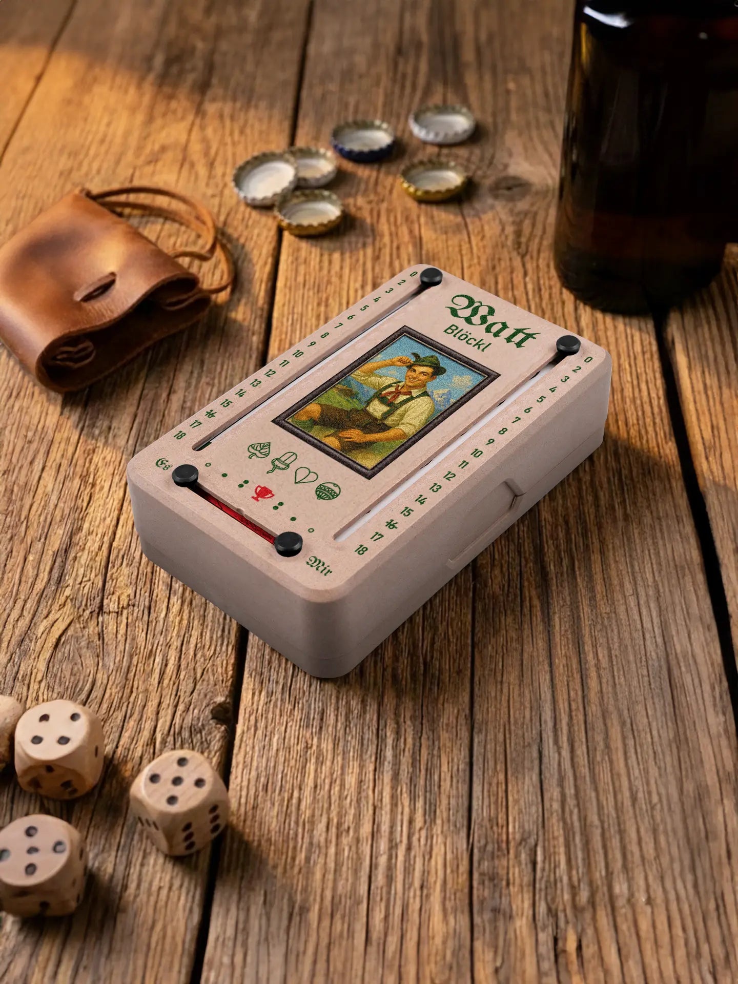 Wattblöckl in Holzoptik mit schwarzen Pins und Alpenbua-Motiv. Platziert auf Holztisch mit Würfeln und Kronkorken.