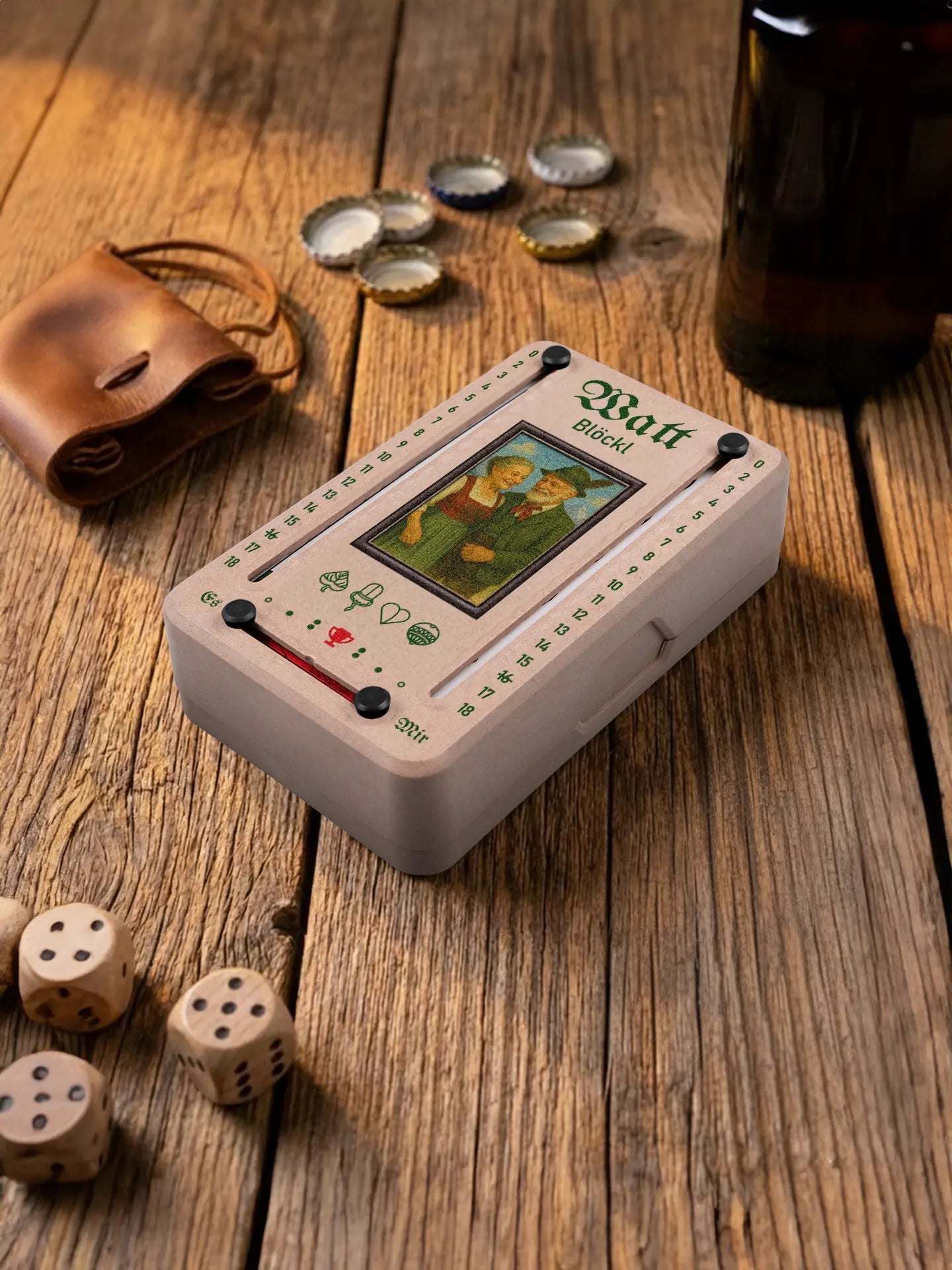 Wattblöckl in Holzoptik mit schwarzen Pins und Alpenliebe-Motiv. Platziert auf Holztisch mit Würfeln und Kronkorken.