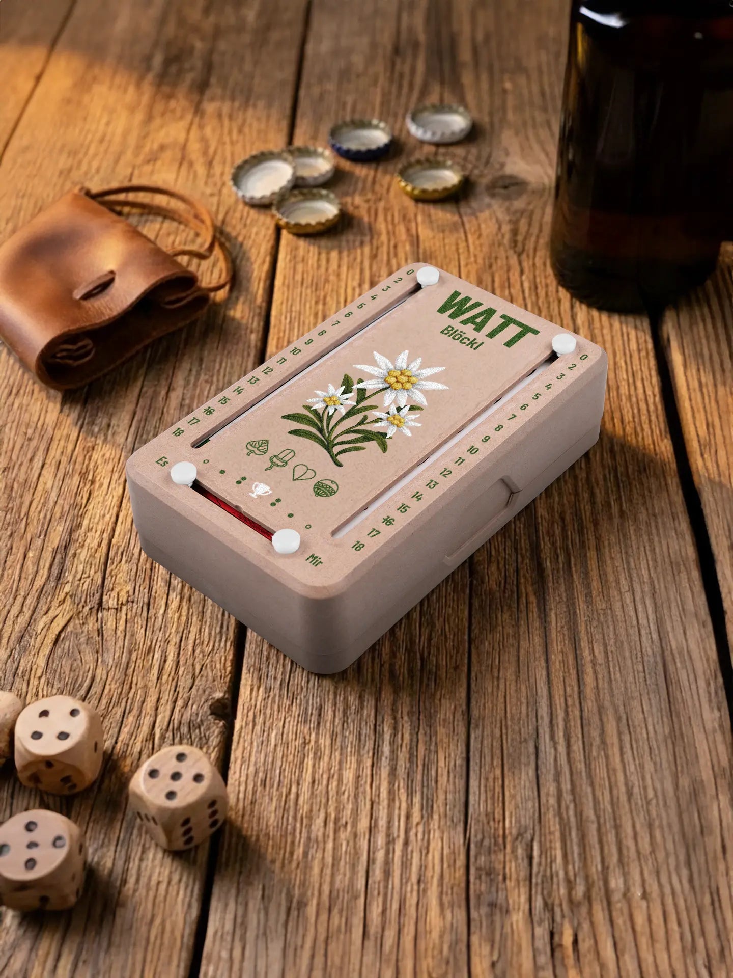 Wattblöckl in Holzoptik mit weissen Pins und Edelweiss-Motiv. Platziert auf Holztisch mit Würfeln und Kronkorken.