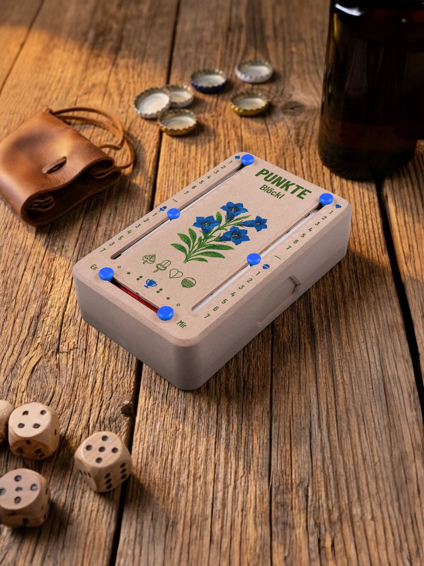 Punkteblöckl in Holzoptik mit blauen Pins und Enzian-Motiv. Platziert auf Holztisch mit Würfeln und Kronkorken.