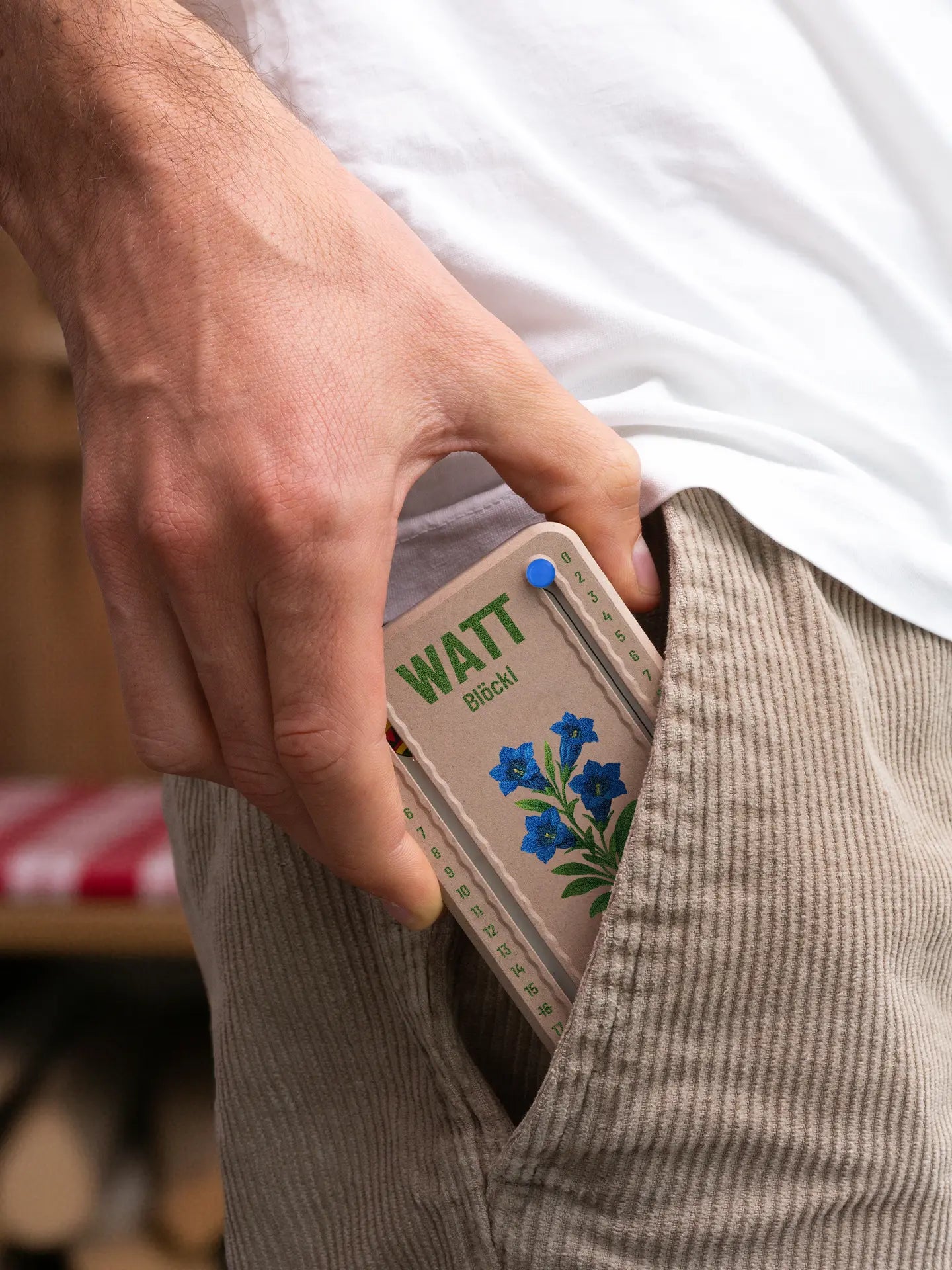 Wattblöckl in Holzoptik mit blauen Pins und Enzian-Motiv wird aus der Hosentasche gezogen.