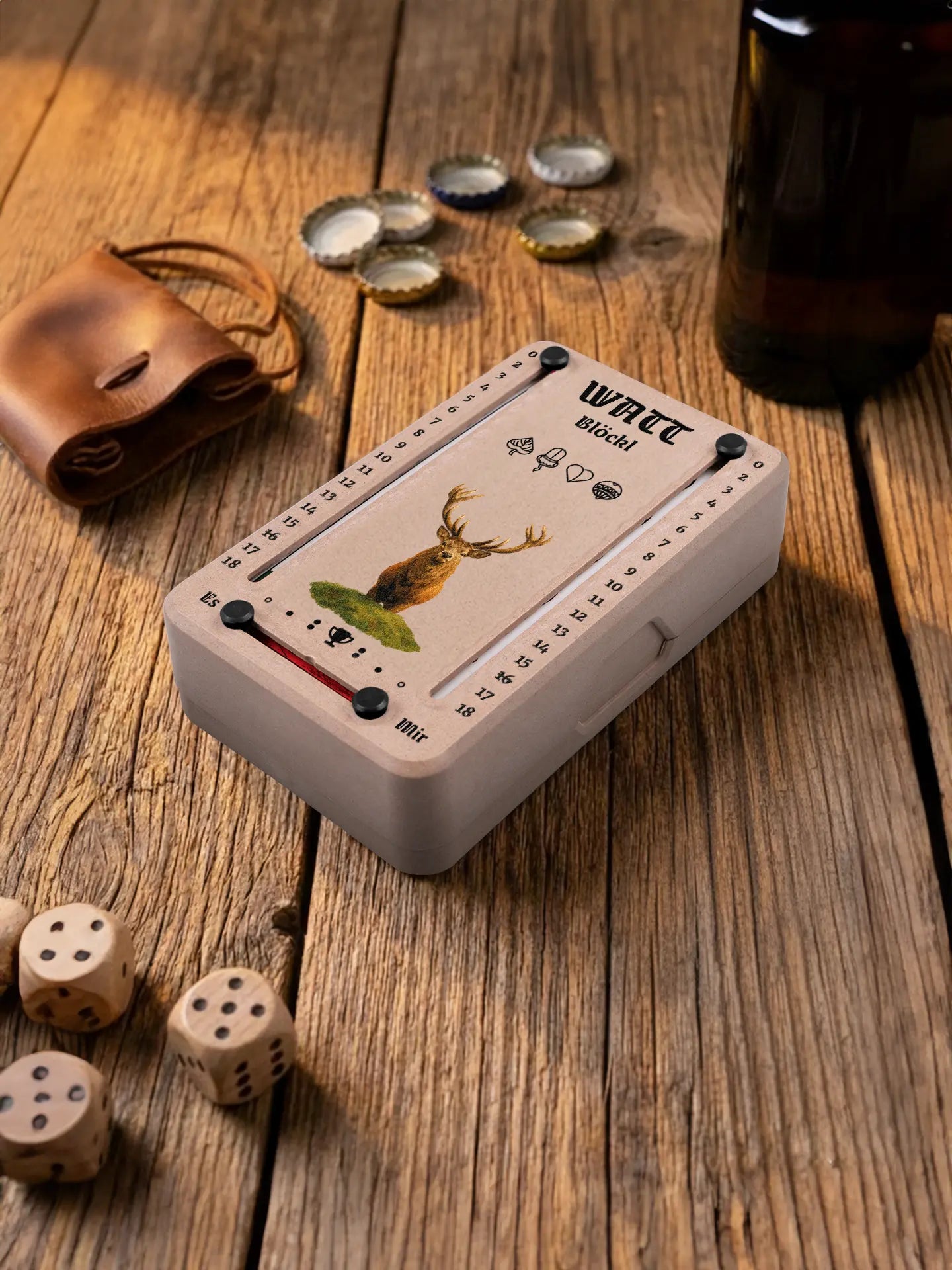 Wattblöckl in Holzoptik mit schwarzen Pins und Hirsch-Motiv. Platziert auf Holztisch mit Würfeln und Kronkorken.