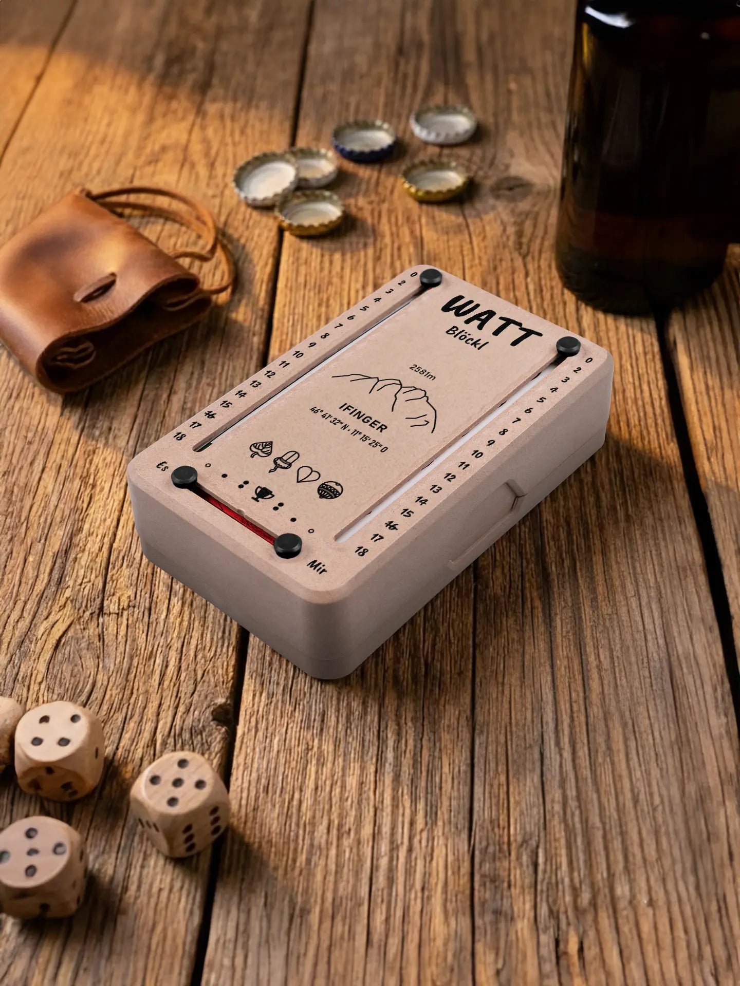Wattblöckl in Holzoptik mit schwarzen Pins und Ifinger-Motiv. Platziert auf Holztisch mit Würfeln und Kronkorken.