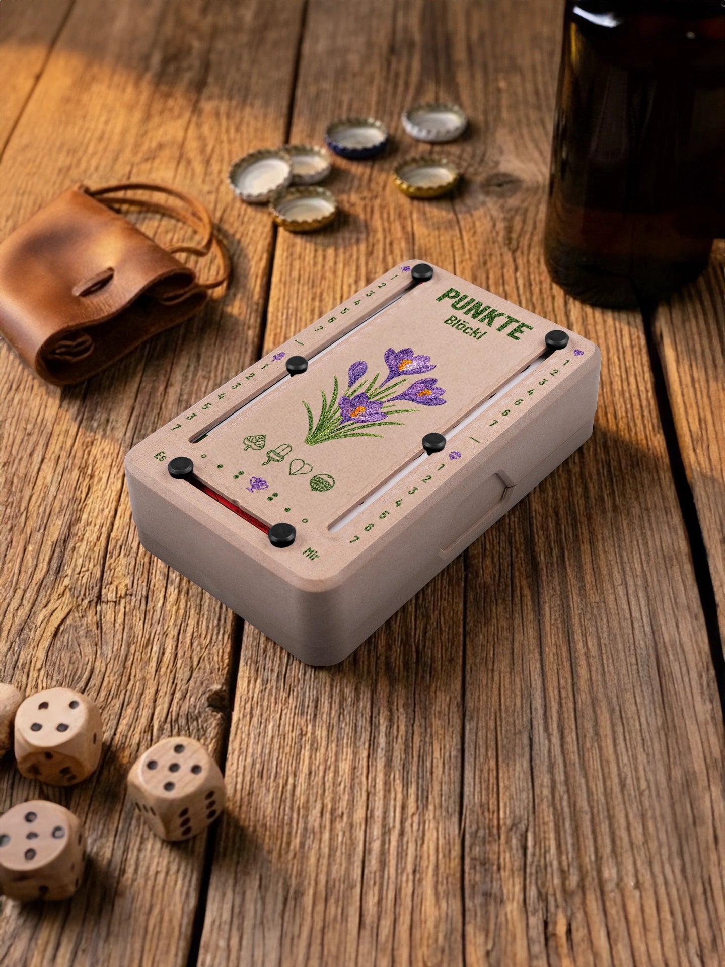 Punkteblöckl in Holzoptik mit schwarzen Pins und Krokus-Motiv. Platziert auf Holztisch mit Würfeln und Kronkorken.