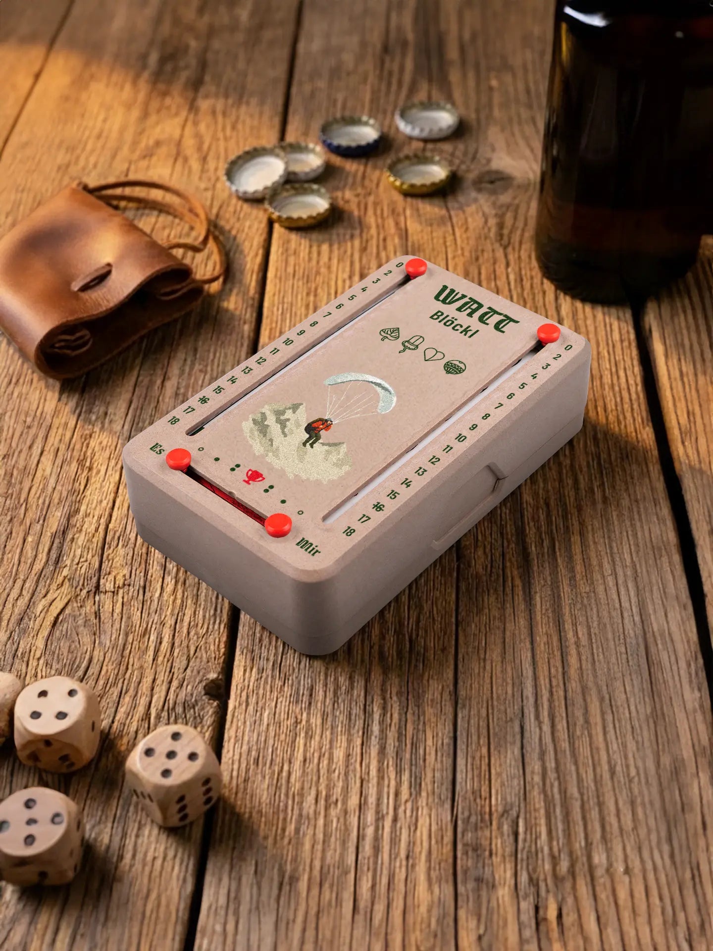 Wattblöckl in Holzoptik mit roten Pins und Paragleiter-Motiv. Platziert auf Holztisch mit Würfeln und Kronkorken.
