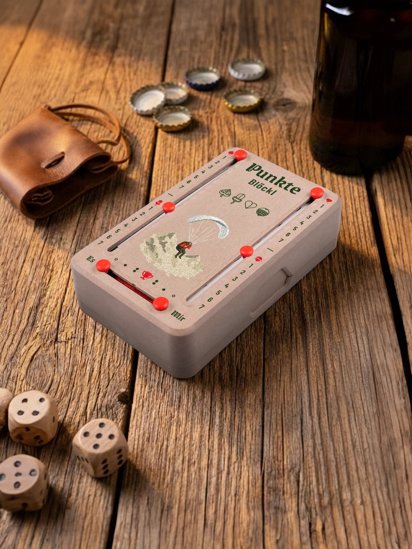 Punkteblöckl in Holzoptik mit roten Pins und Paragleiter-Motiv. Platziert auf Holztisch mit Würfeln und Kronkorken.