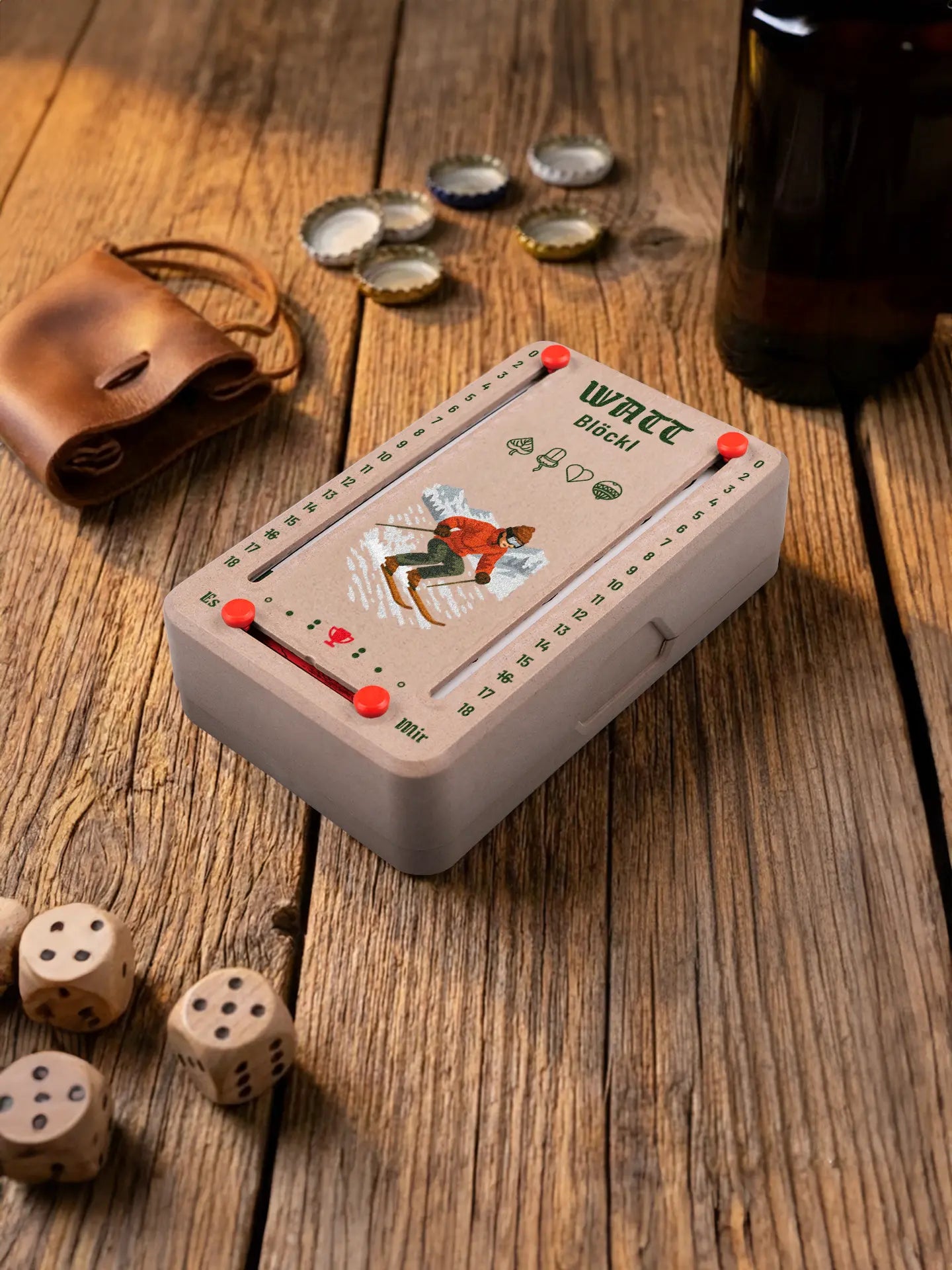 Wattblöckl in Holzoptik mit roten Pins und Skifahrer-Motiv. Platziert auf Holztisch mit Würfeln und Kronkorken.