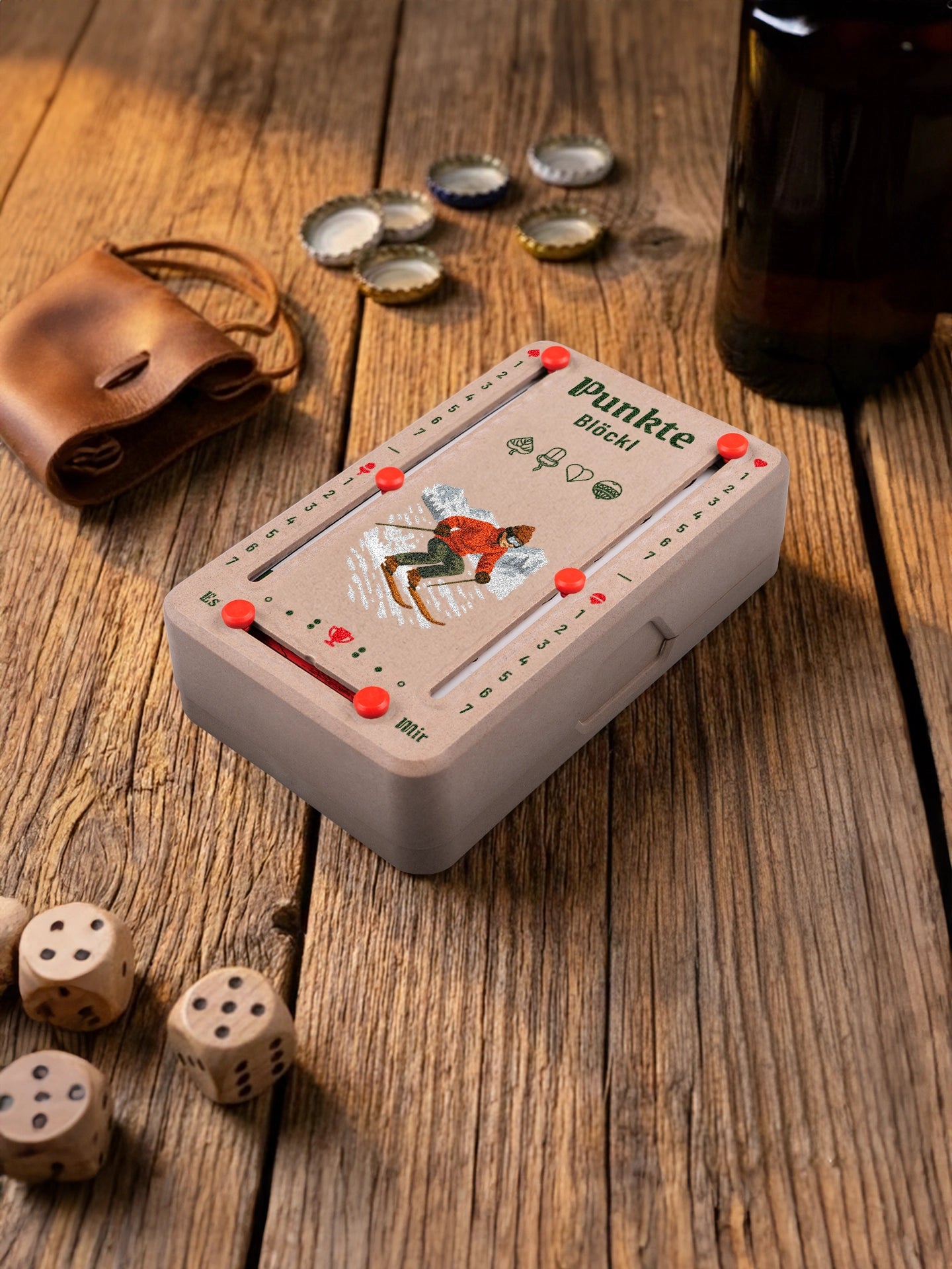 Punkteblöckl in Holzoptik mit roten Pins und Skifahrer-Motiv. Platziert auf Holztisch mit Würfeln und Kronkorken.
