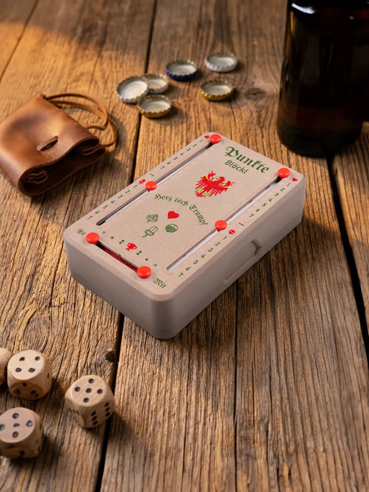 Punkteblöckl in Holzoptik mit roten Pins und Suedtirol-Motiv. Platziert auf Holztisch mit Würfeln und Kronkorken.