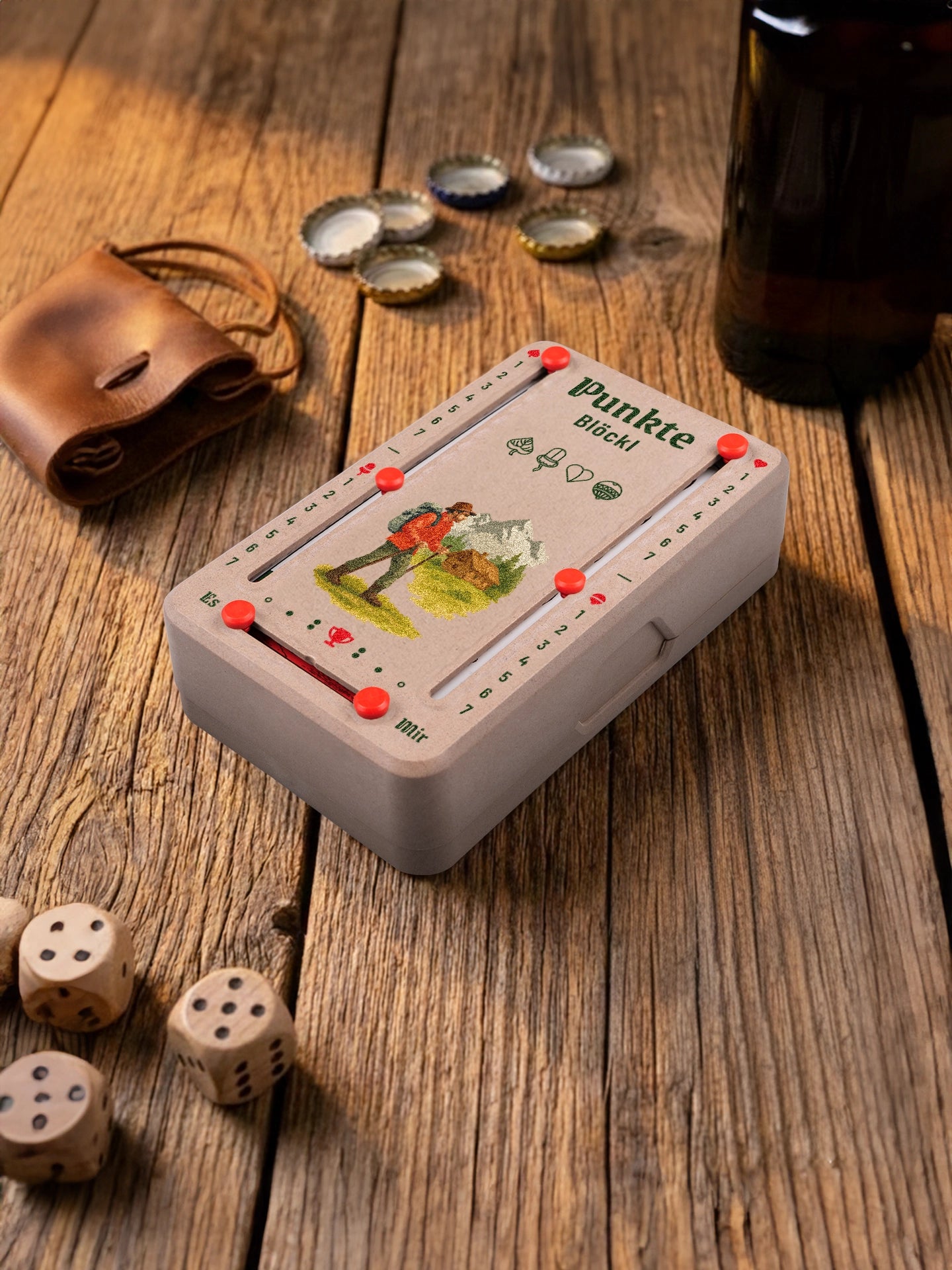 Punkteblöckl in Holzoptik mit roten Pins und Wanderer-Motiv. Platziert auf Holztisch mit Würfeln und Kronkorken.