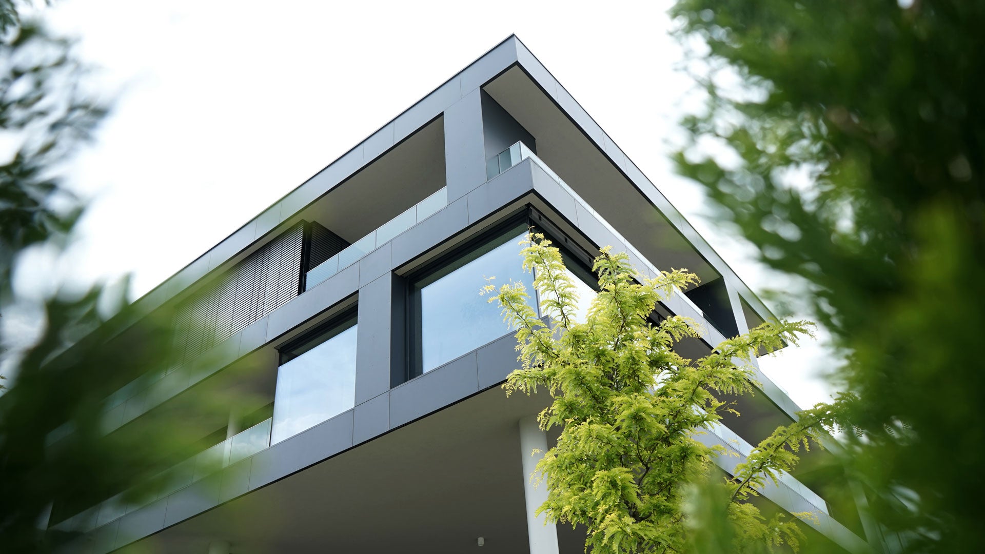 Firmengebäude mit Pflanzen und Bäumen in Vordergrund. Himmel ist bewölkt.